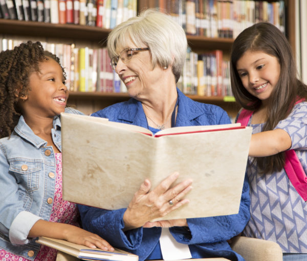 Senior adult teacA senior female volunteer reads a book to elementary students in school classroom or library setting.